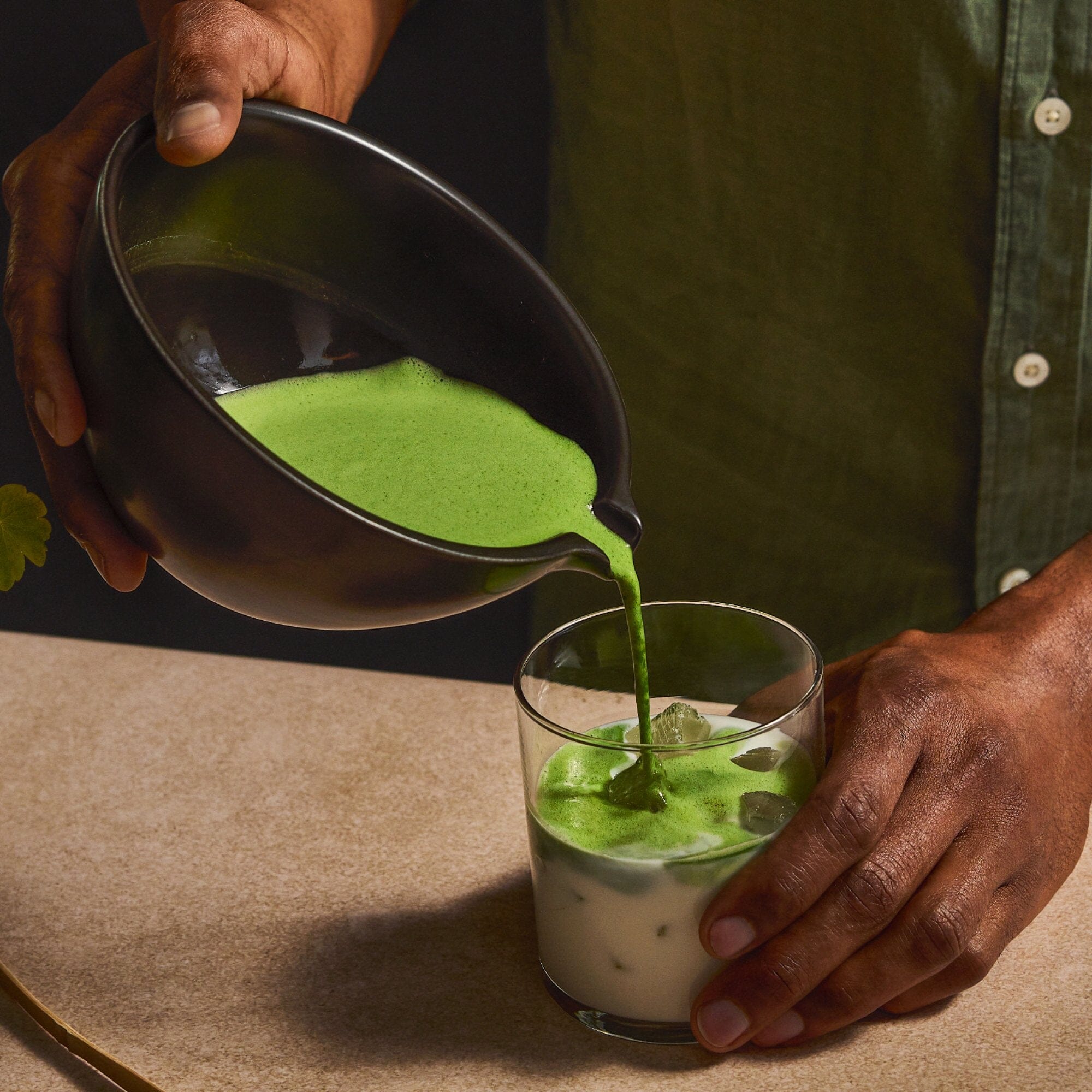 A person pours a vibrant green Rishi Tea & Botanicals Barista Matcha mixture from a black bowl into a glass of iced milk on a countertop.