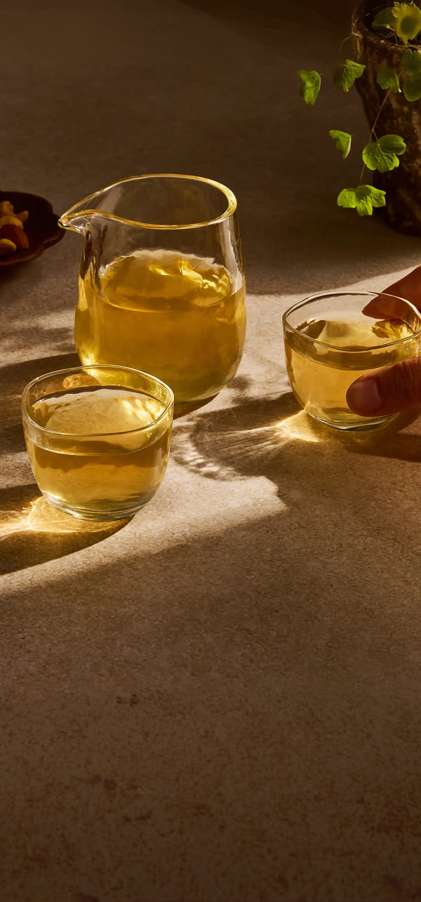 A clear glass pitcher and two small glasses filled with golden yellow tea, arranged on a warm beige surface under dramatic sunlight and shadows.