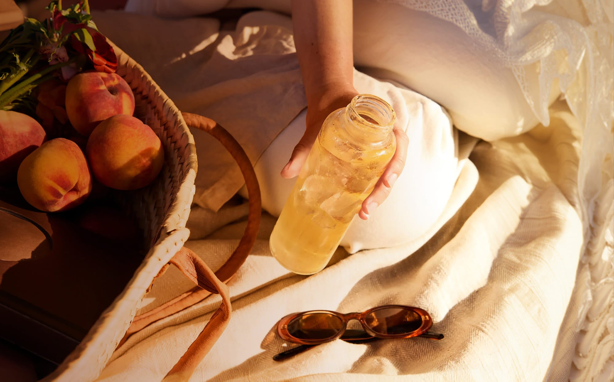 A person in a white dress holds a clear glass bottle filled with a yellow drink on a cream fabric surface. A woven basket with peaches, sunglasses, and flowers appears at the left edge in warm, sunlit styling.