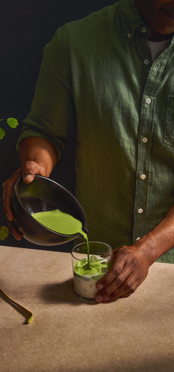 A person in a green button-up shirt pours bright green liquid from a metal bowl into a glass on a beige tabletop against a dark background.