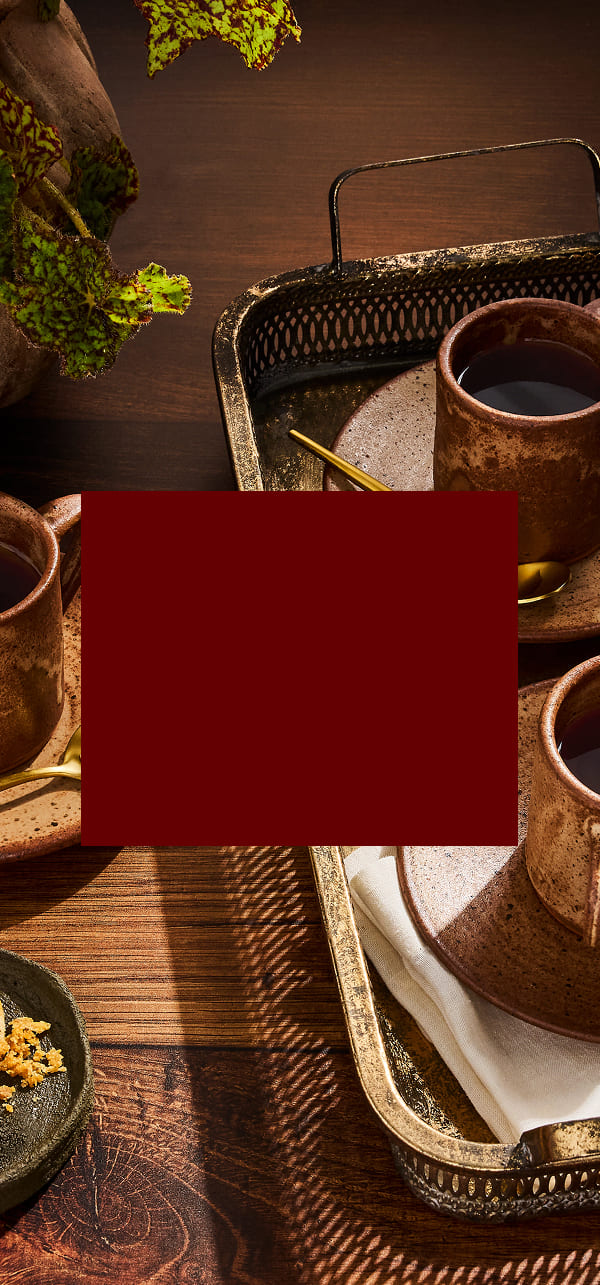 A styled tabletop scene with several rustic brown ceramic cups and saucers on metal trays over a dark wooden table, plus a large solid dark red square obscuring the center.