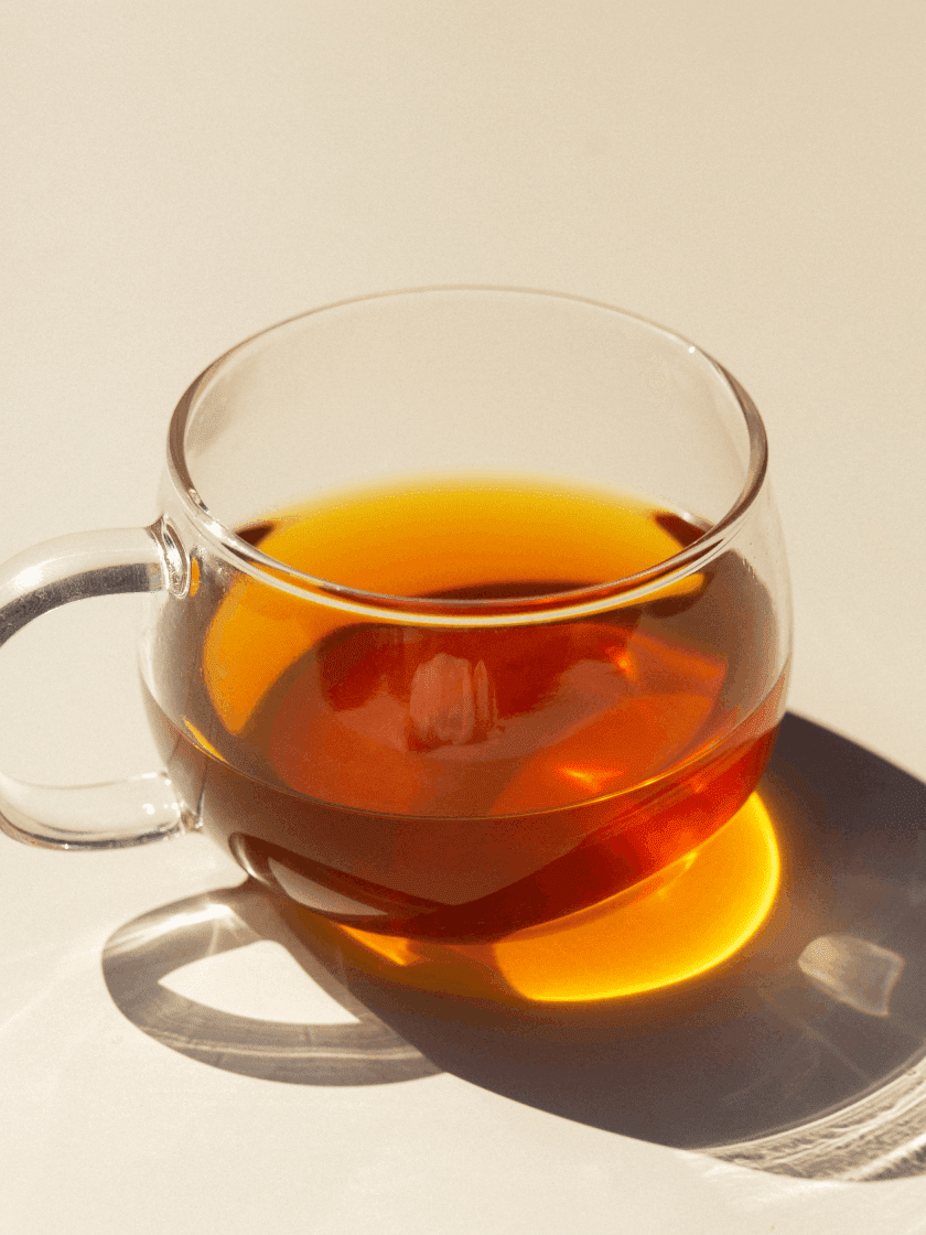 A clear glass cup filled with red liquid, a hand stirring with a cinnamon stick, and a whole plum placed beside the cup.