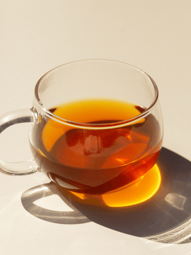 A clear glass cup filled with red liquid, a hand stirring with a cinnamon stick, and a whole plum placed beside the cup.