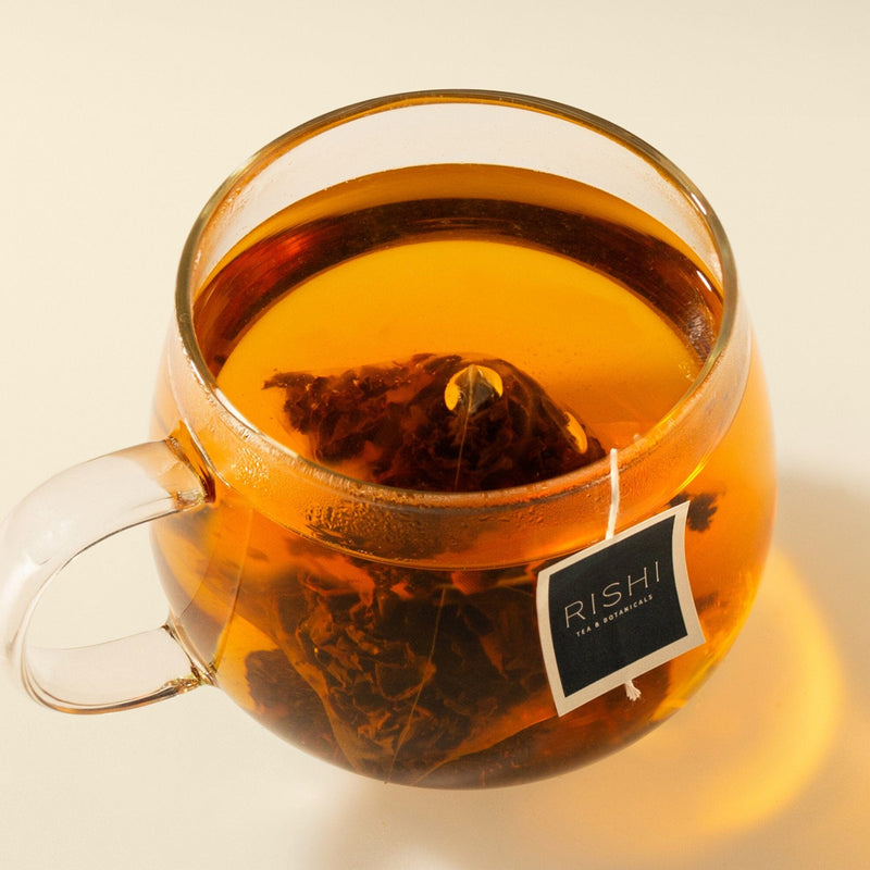 A clear glass mug filled with amber-hued tea displays a submerged tea bag, its string adorned with a visible Rishi Tea & Botanicals tag, suggesting an infusion of red rooibos and delicate rose petals.