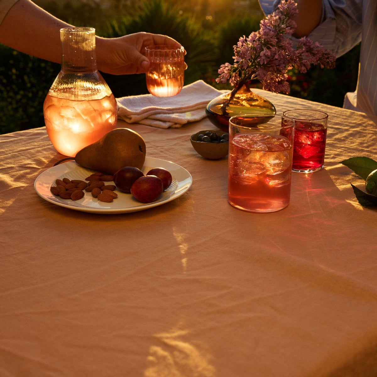 A sunlit outdoor table is set with drinks from Rishi Tea & Botanicals—perhaps an herbal blend or rooibos tea—a plate of almonds, grapes, and a pear, a small bowl of olives, and a vase with lilac flowers. Two people are holding glasses.
