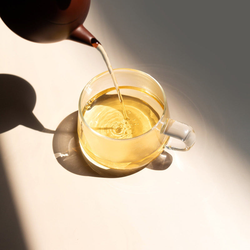 A glass cup is being filled with Golden Peony oolong tea by Rishi Tea & Botanicals from a brown teapot on a white surface, sunlight highlighting the tea's light color and subtle orchid aromatics.