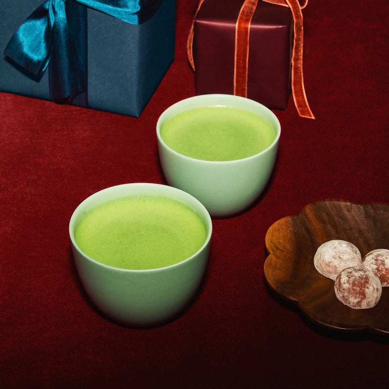 Two light green cups filled with matcha tea sit on a red surface next to a wooden plate with three chocolate truffles and two gift boxes wrapped with ribbons.