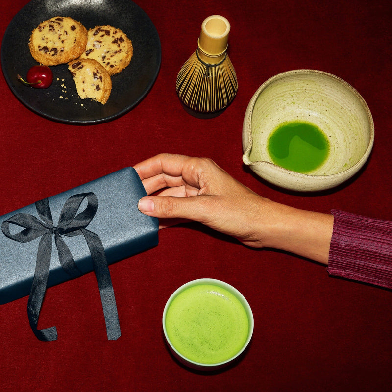 A hand reaches for a blue gift box on a red surface next to a bowl and cup of matcha, a whisk, and a plate with cookies and a cherry.