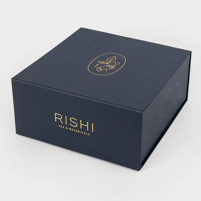 A dark blue box with gold embossed “RISHI TEA & BOTANICALS” text and a minimalist leaf logo on the lid, set against a plain white background.