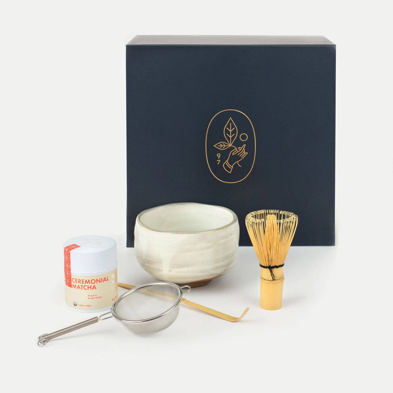 Ceremonial matcha kit with a navy box, matcha tin, white bowl, bamboo whisk, metal sifter, and bamboo scoop arranged on a white background.