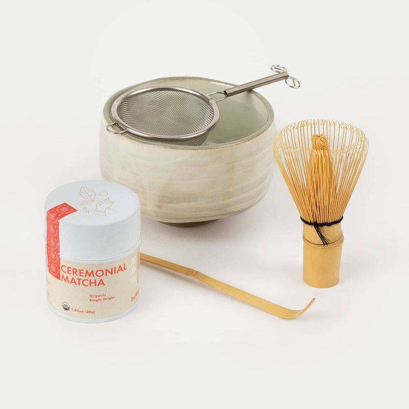 Ceremonial matcha tea set with a tin of matcha powder, a ceramic bowl, a metal sifter, a bamboo whisk, and a bamboo scoop, all arranged on a white background.