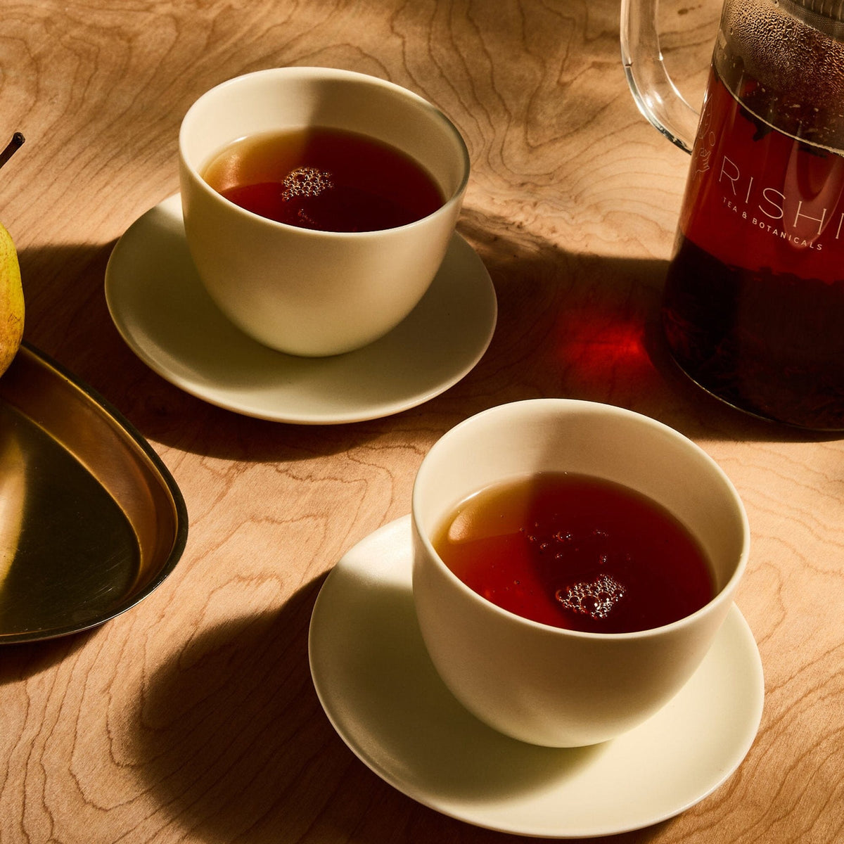 Two white cups filled with tea on saucers sit on a wooden table beside a tea carafe and a metal tray with a pear, the inviting aroma of hot Rishi Tea & Botanicals ginger and tulsi holy basil mingling in the air.