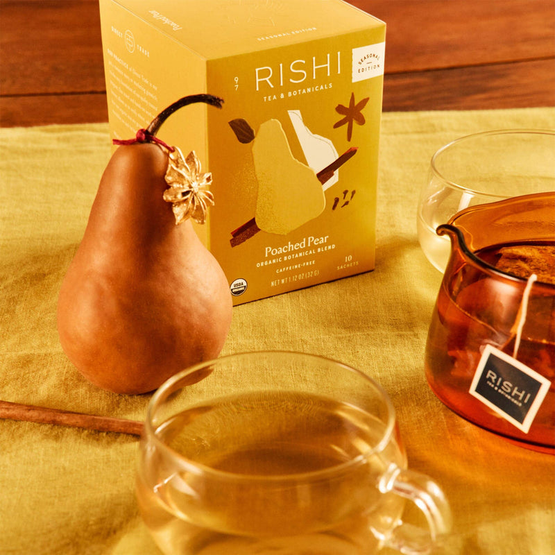 A box of Rishi Tea & Botanicals Poached Pear organic tea is shown with a fresh pear, a cinnamon stick, two glass cups of tea, and a glass teapot on a yellow cloth.