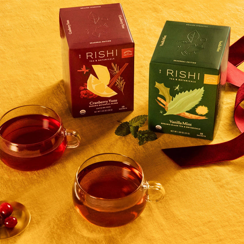 Two glass cups of seasonal tea are on a yellow surface beside Rishi Tea & Botanicals boxes labeled 