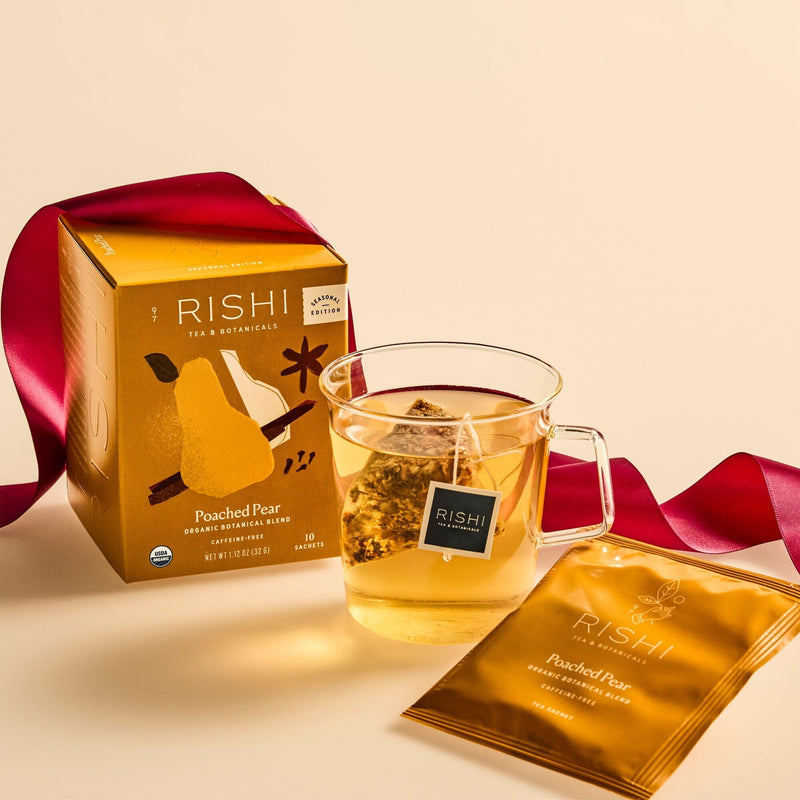 A box of Rishi Tea & Botanicals Poached Pear tea, a glass mug of tea, a tea bag, and a red ribbon rest on a beige surface—showcasing the blend’s delicate pear essence and subtle holiday spice notes.