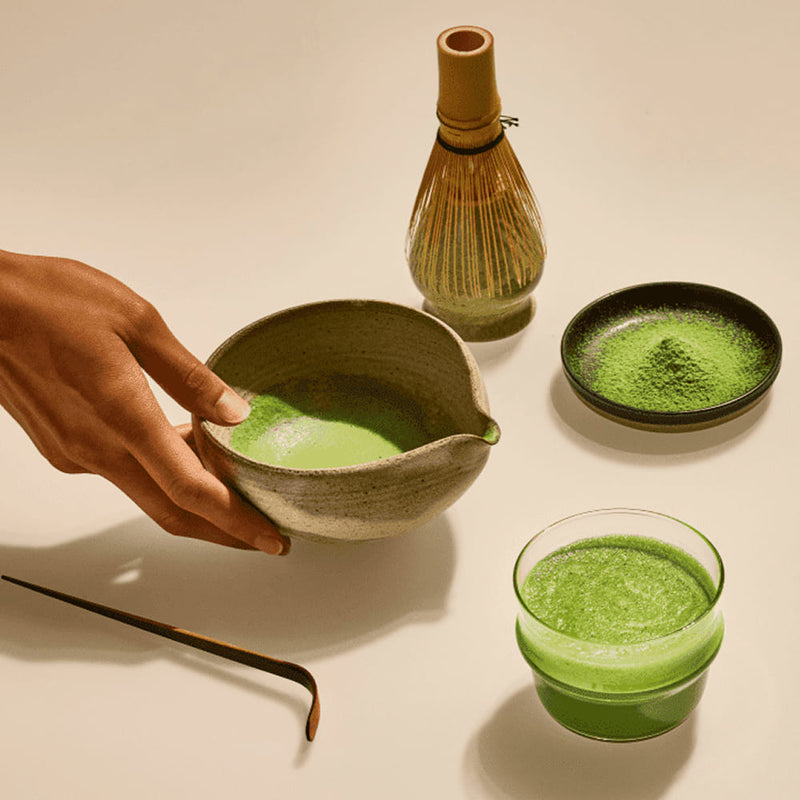 A hand holds a small ceramic bowl filled with bright green matcha powder or tea, with a bamboo whisk, a small dish of loose green powder, and a glass of green beverage arranged on a warm cream background.