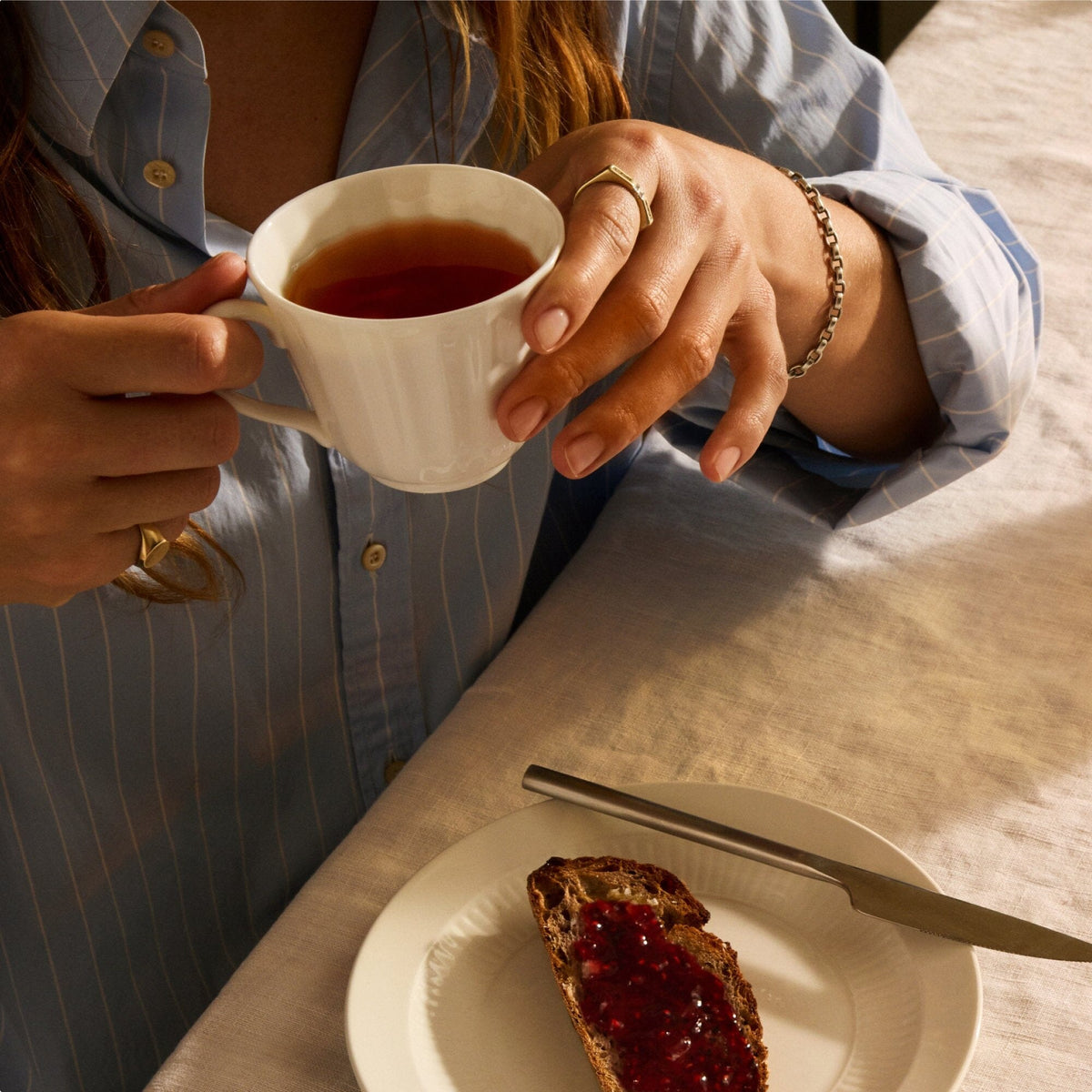 A person in a blue shirt holds a cup of Rishi Tea & Botanicals at a table with a plate of bread topped with red jam and a butter knife.