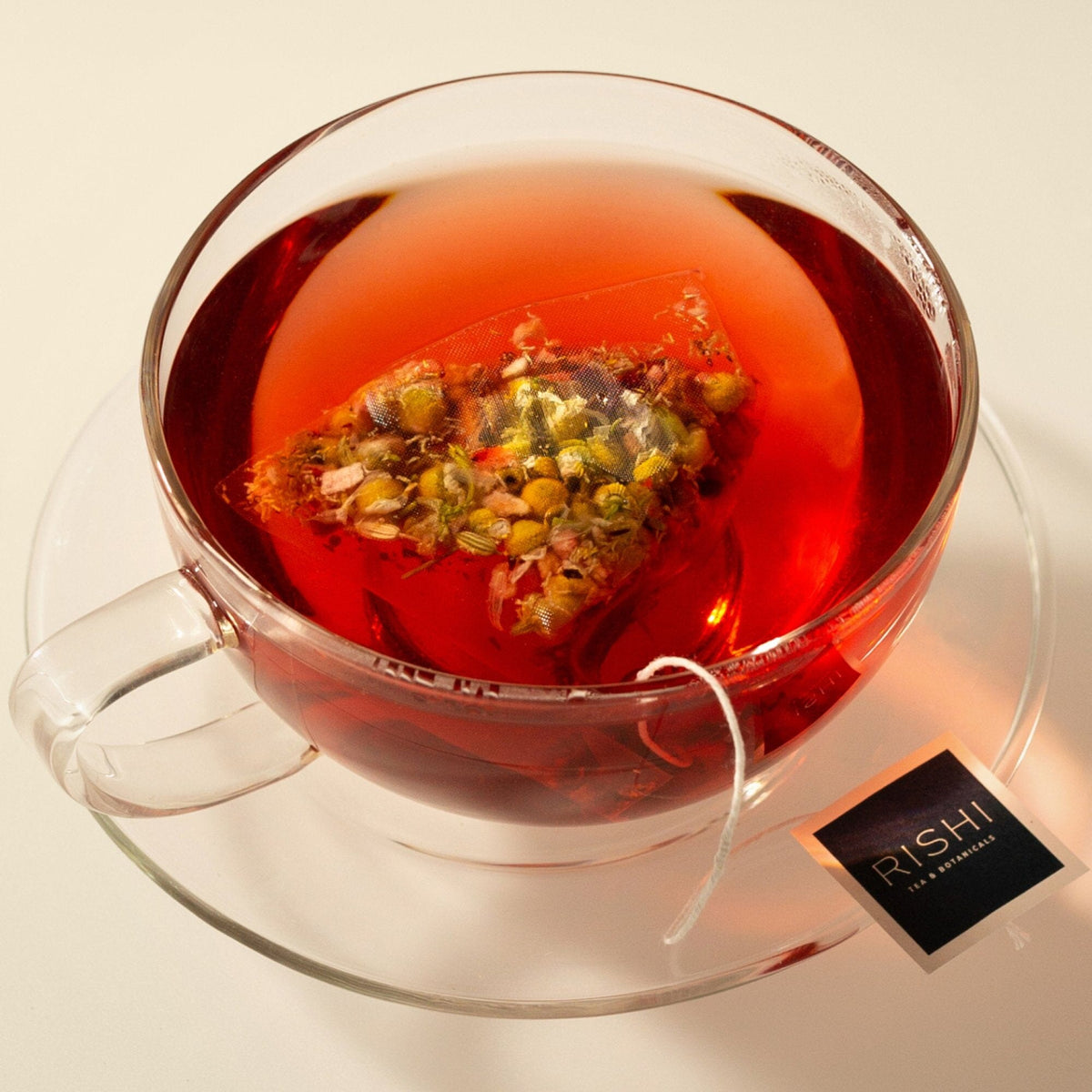 A clear glass cup of red herbal tea, featuring a chamomile tea bag from Rishi Tea & Botanicals, sits atop a glass saucer. The vibrant hue suggests a flavorful infusion of hibiscus tea, with the tea bag label marked "RISHI.
