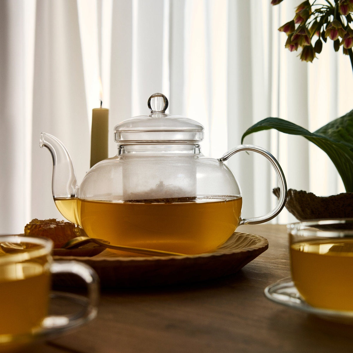 A glass teapot from Rishi Tea & Botanicals, filled with tea, rests on a wooden tray alongside two glass teacups, a lit candle, and a potted plant atop a wooden table, emanating the invigorating aroma of energizing ginger.