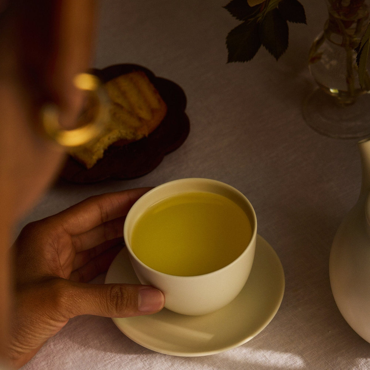 A person holds a white cup of golden Rishi Tea & Botanicals chamomile tea on a saucer, with cake and flowers in the background.