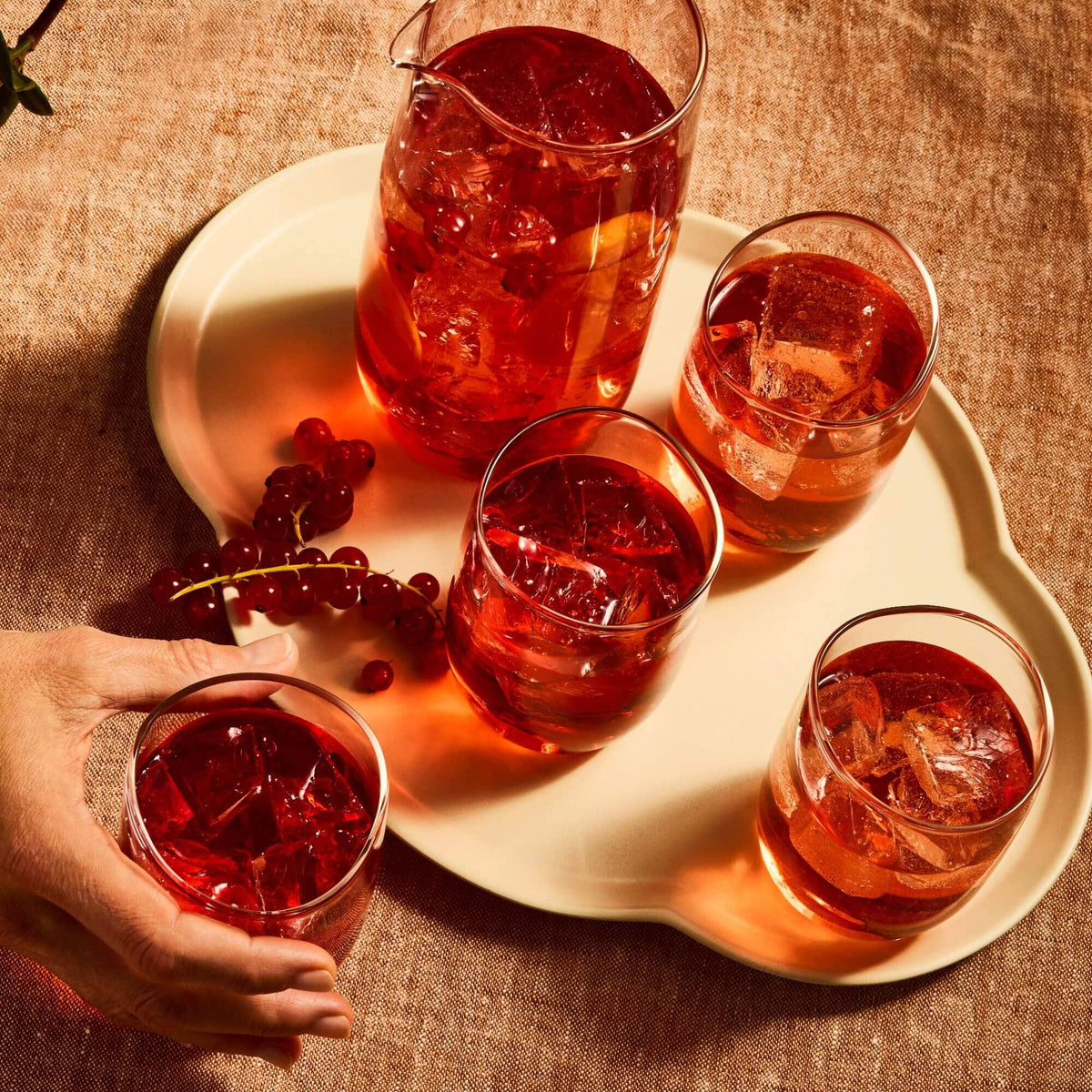 A white tray holds a pitcher and four glasses of Rishi Tea & Botanicals Hibiscus Berry sangria garnished with red currants, as a hand reaches for one glass.