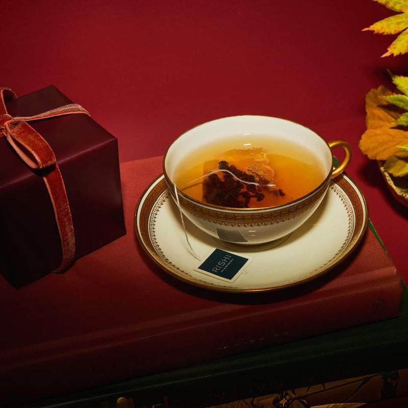 A cup of tea with a teabag sits on a saucer next to a wrapped gift box and autumn leaves, all arranged on top of a closed book against a red background.