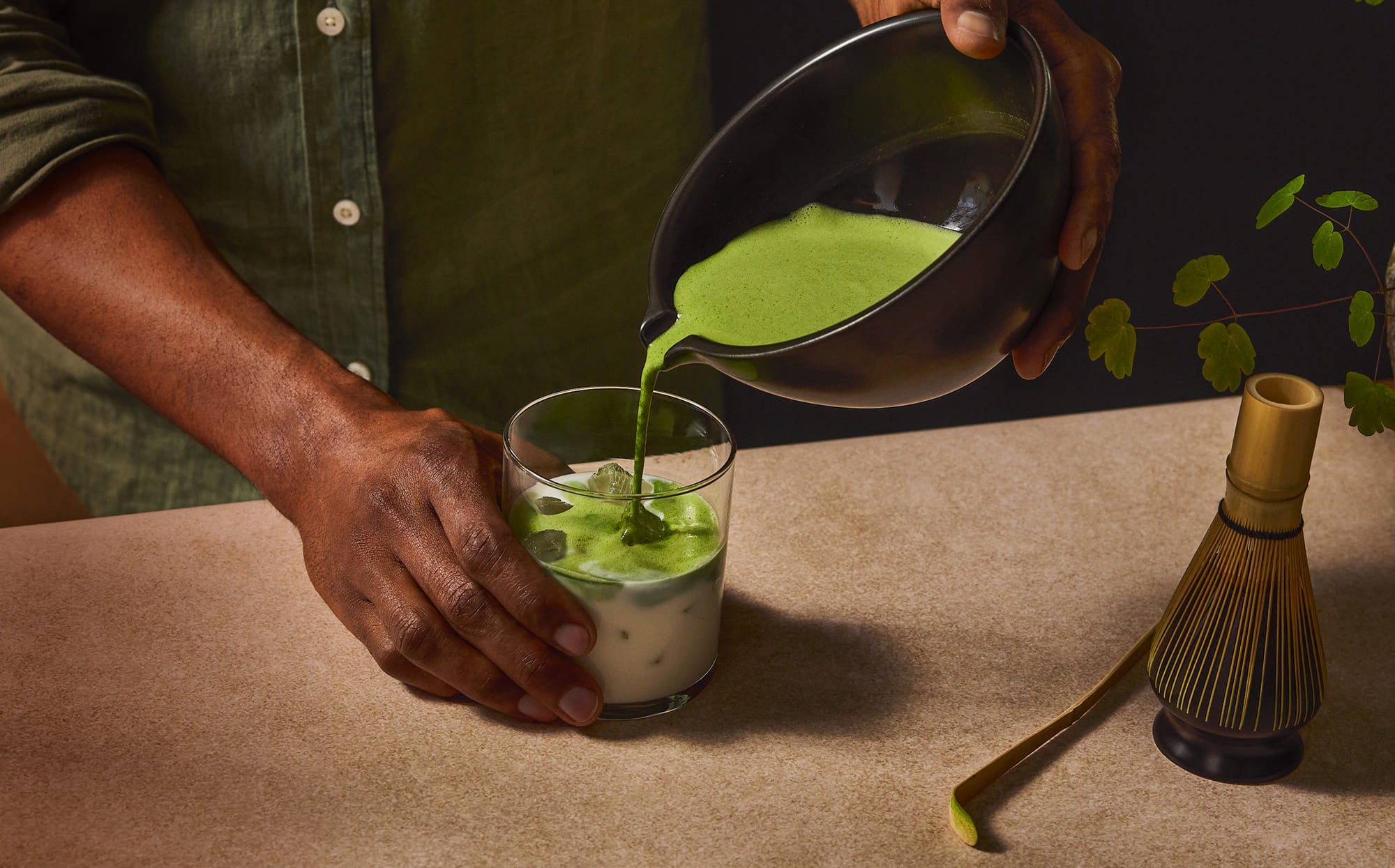 A person pouring bright green matcha tea from a black bowl into a glass of milk and matcha, set on a beige tabletop with a bamboo whisk nearby.
