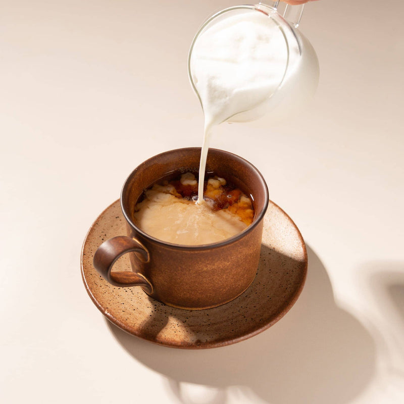 A brown ceramic mug with a matching saucer, filled with a layered drink, while milk is being poured from a clear glass pitcher. The scene is shot from a high angle on a warm beige background with soft shadows.