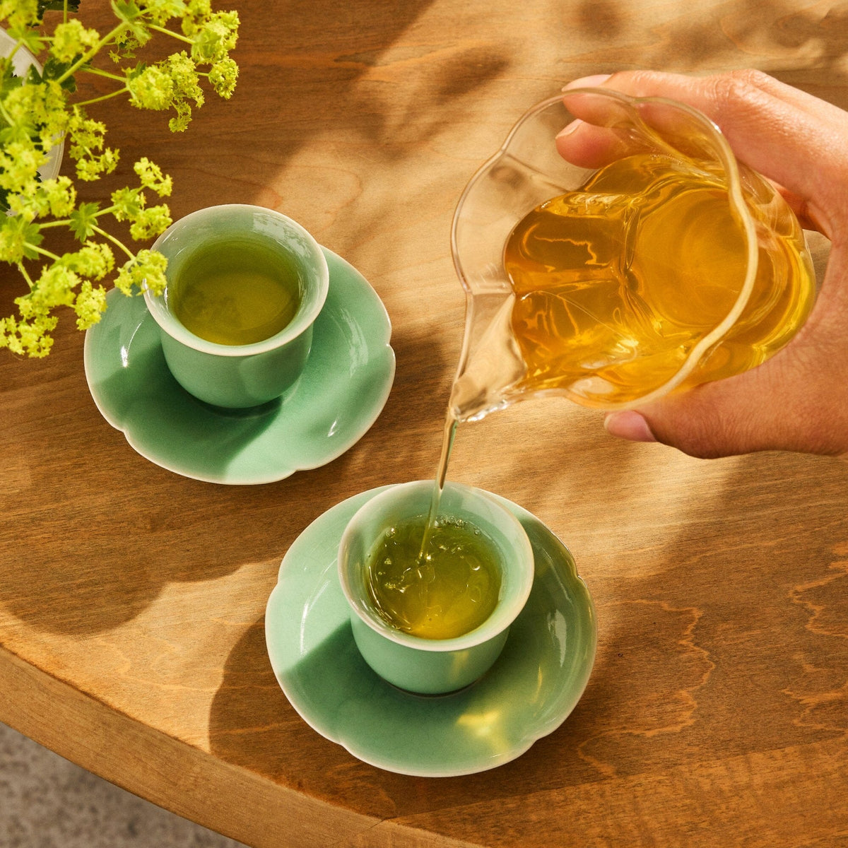 A hand pours Rishi Tea & Botanicals Jasmine Green tea from a glass pitcher into a small green cup on a saucer, beside another filled cup, on a wooden table with green flowers—evoking the spirit of Hubei's Xuan En region.