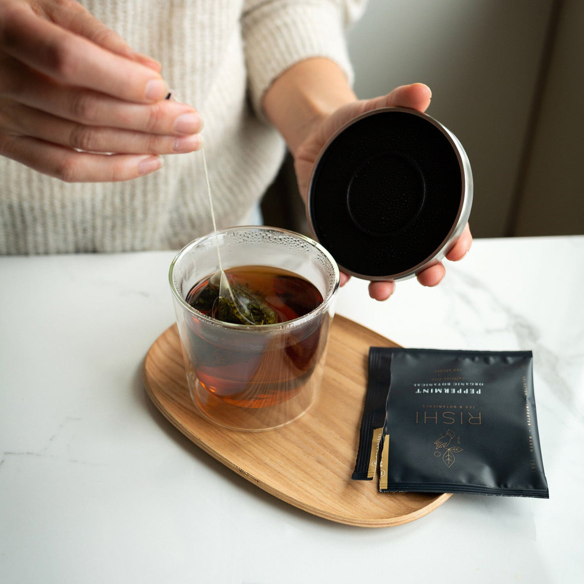 A person places a tea bag in a Kinto Double Wall Glass Cup & Lid, set on a wooden tray with the lid and tea packet, on the marble countertop.
