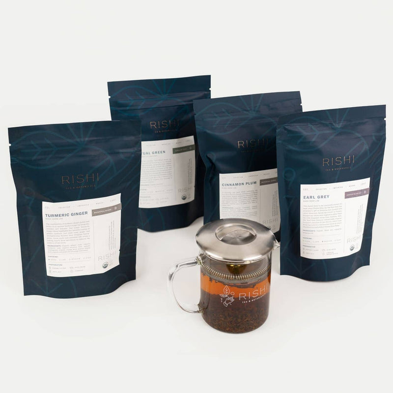 Four Rishi tea bags—Turmeric Ginger, Sencha, Cinnamon Plum, and Earl Grey—stand behind a glass teapot filled with tea, capturing the essence of a curated tea gift box.