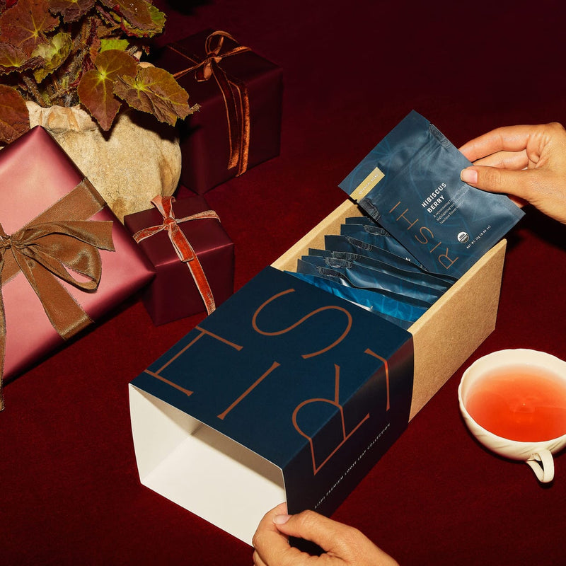 A person pulls a blue packet from a box labeled “RISHI” beside wrapped gifts, a potted plant, and a cup of tea on a red surface.