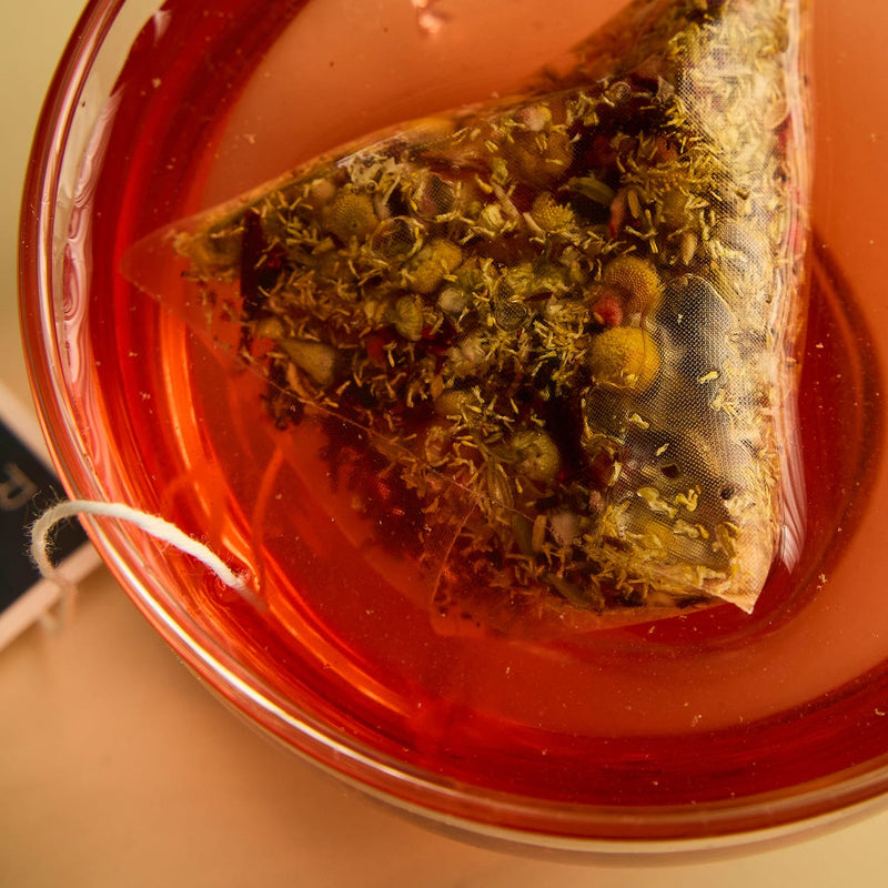 A pyramid tea bag steeps in a cup of reddish herbal tea, with loose herbs visible inside the mesh bag.