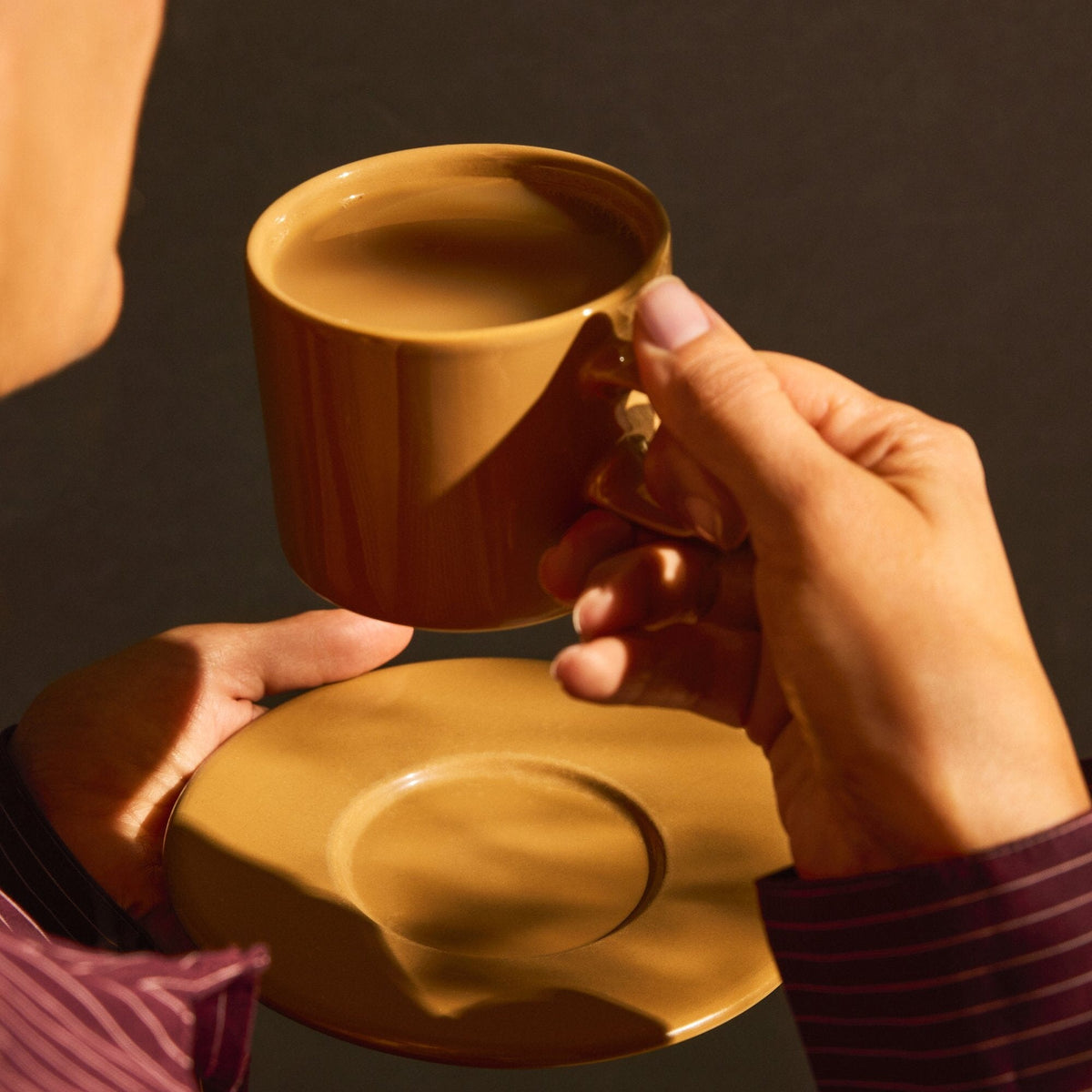 A person holds a brown cup of Rishi Tea & Botanicals Masala Chai, infused with quality spices, in one hand and a matching saucer in the other against a dark background.
