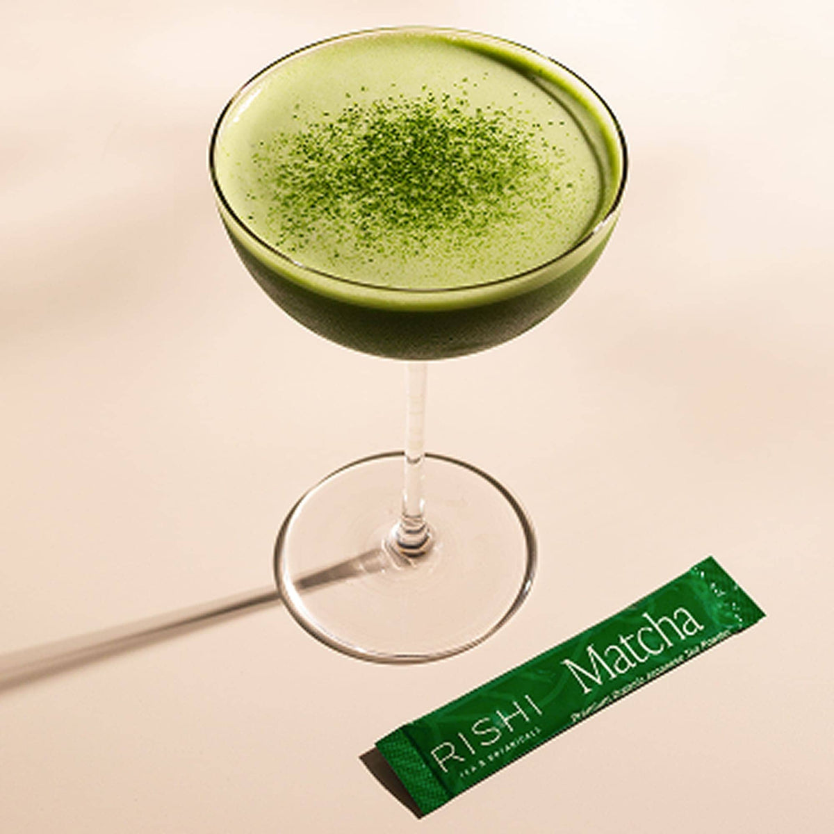 A glass of green Japanese matcha drink topped with matcha powder sits beside a single-serve stick packet from Rishi Tea & Botanicals on a light surface.
