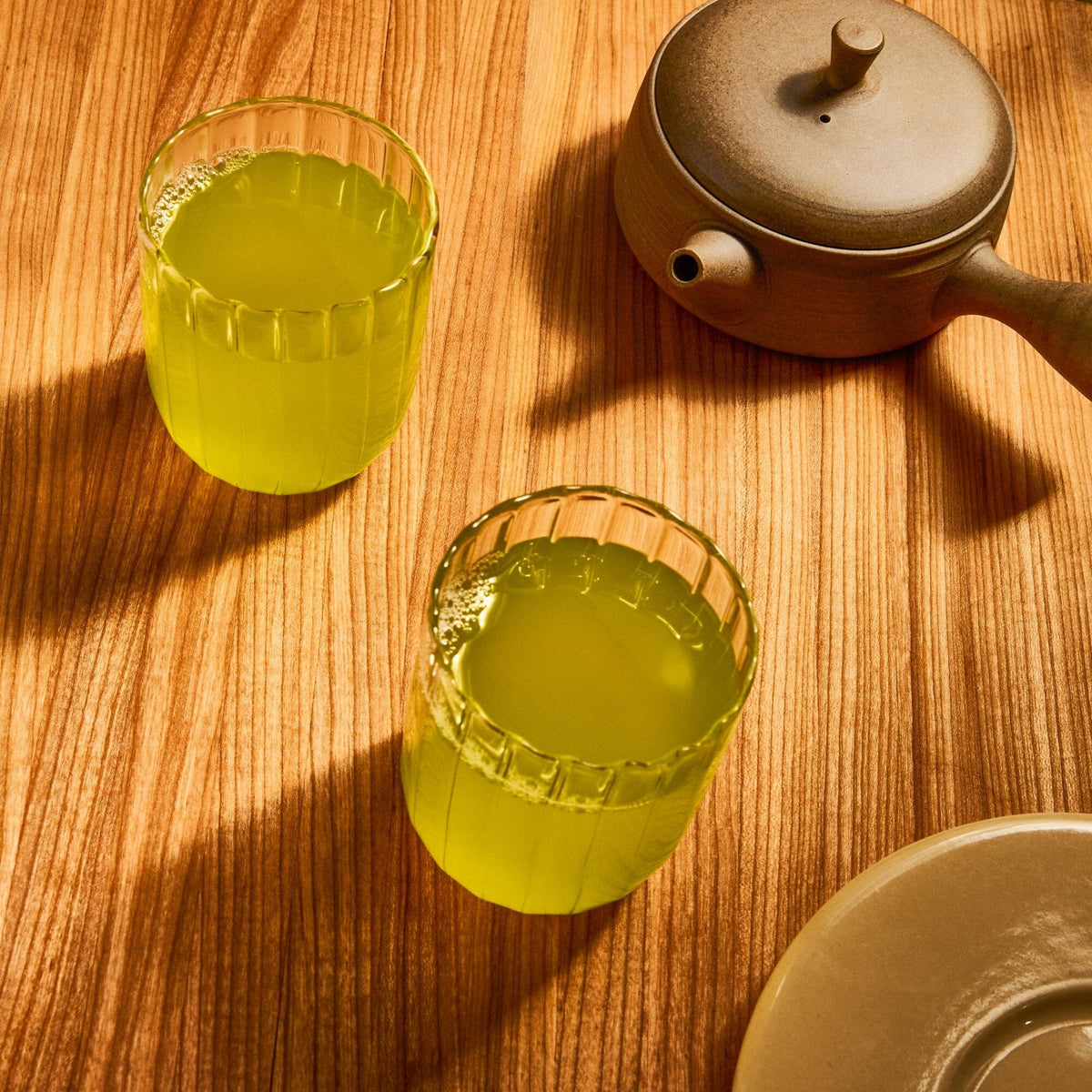 Two glasses of matcha green tea by Rishi Tea & Botanicals on a wooden surface beside a ceramic teapot and plate, sunlight casting gentle shadows across the scene.