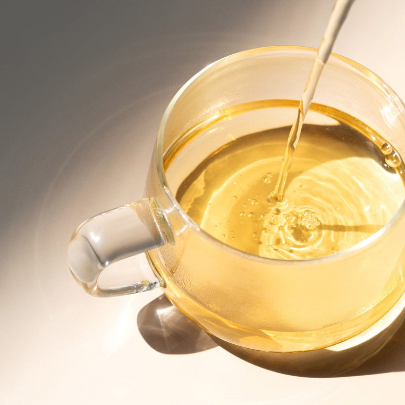 A clear glass cup is being filled with light yellow Golden Peony oolong tea from Rishi Tea & Botanicals, as sunlight creates elegant reflections and shadows on the surface.