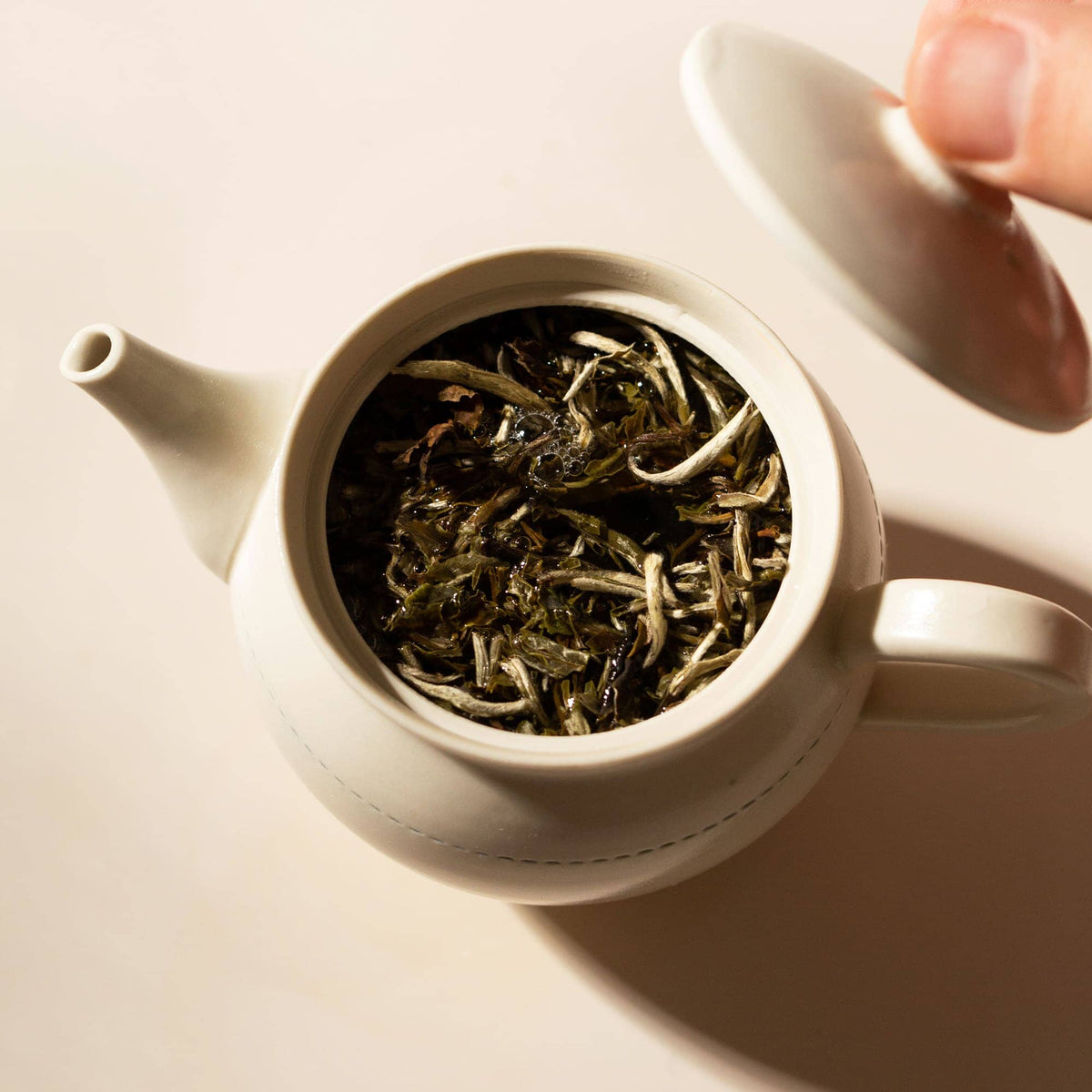 White ceramic teapot with green tea leaves on a beige background