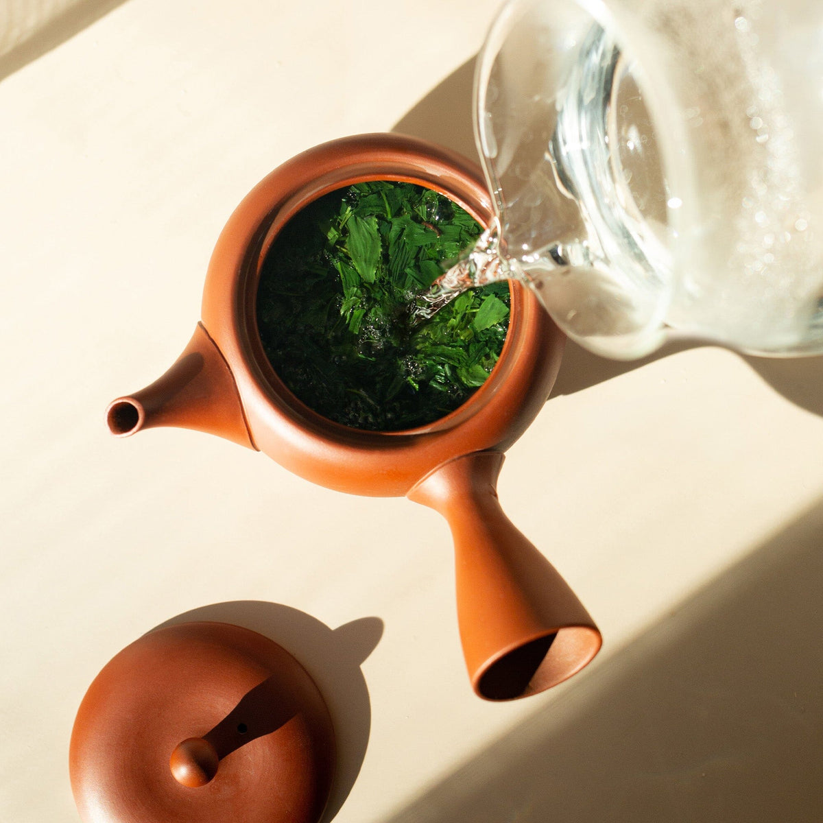 A glass pitcher pours hot water into a brown teapot filled with Rishi Tea & Botanicals organic kuradashi gyokuro leaves, the lid resting nearby on a light surface.