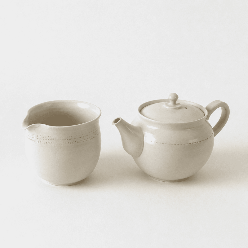 A beige ceramic milk jug and kyusu teapot from Rishi Tea & Botanicals sit side by side on a plain white background, forming a minimalist collectible teaware set.