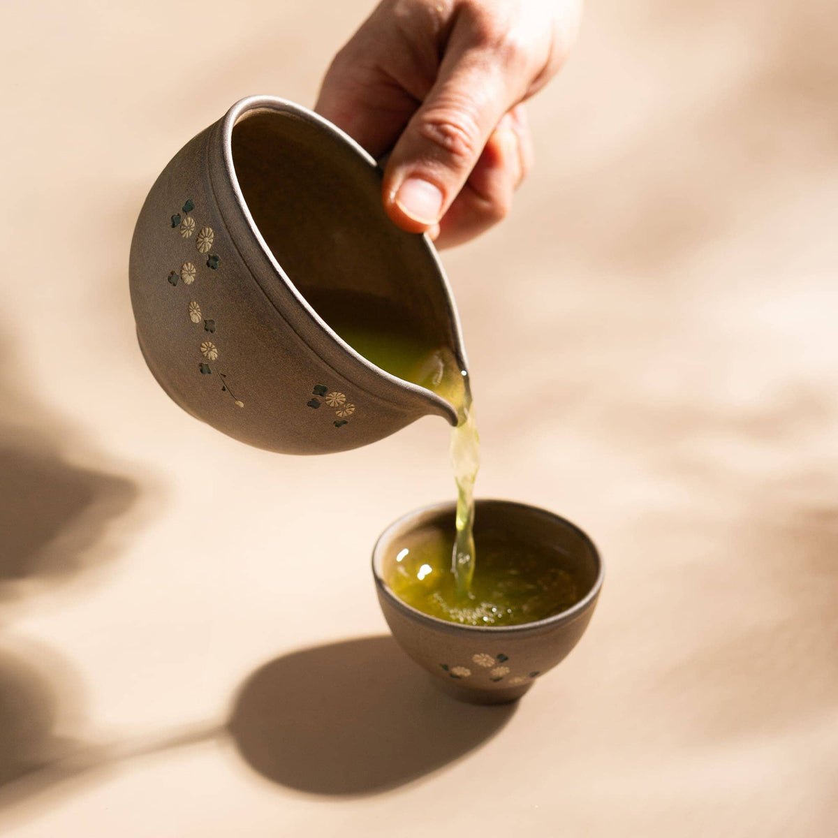 A hand pours green tea from a Chato Co., Ltd. Tokoname shudei clay teapot into a matching teacup—both collectible vessels—on a neutral surface.