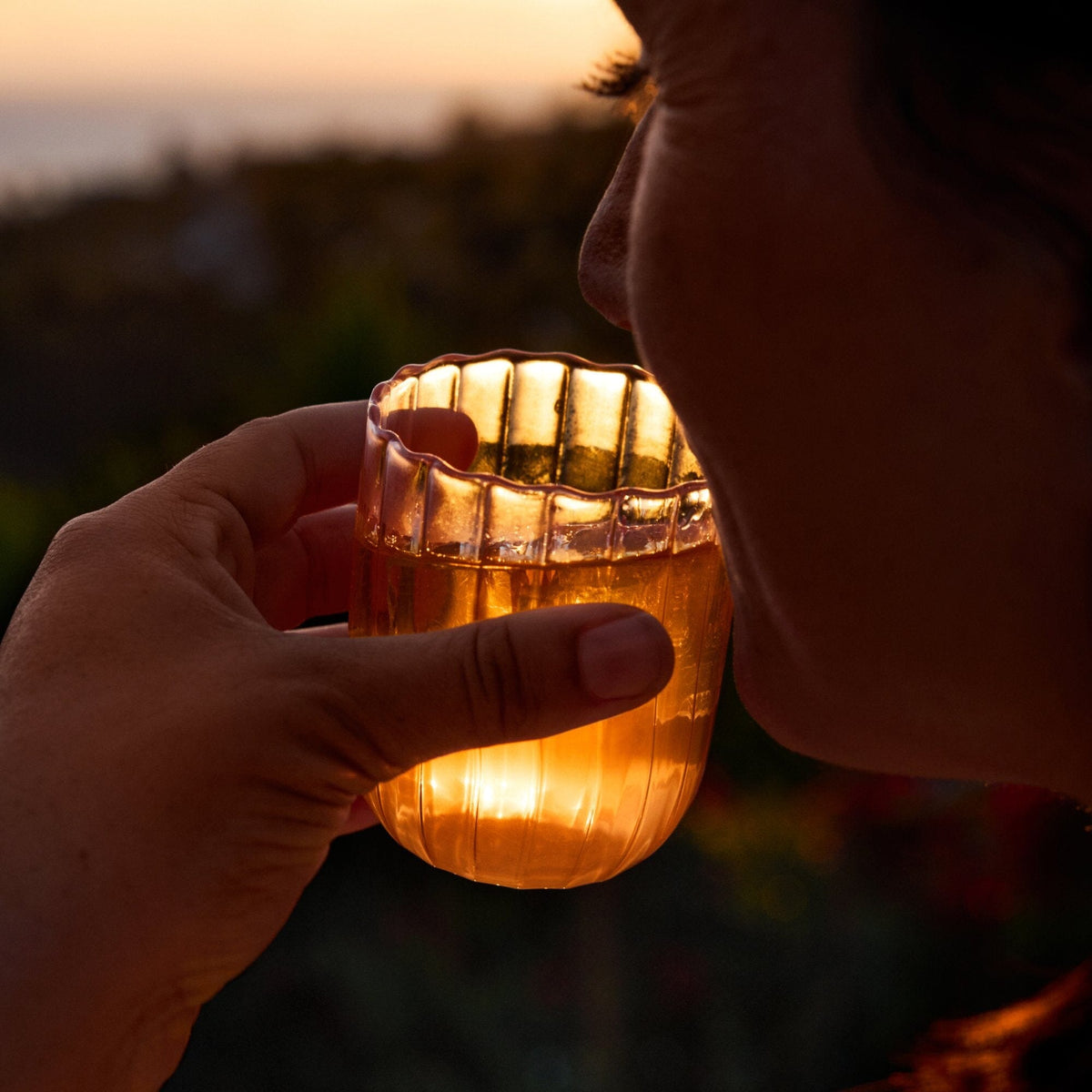 A person holds a glass of amber-colored Rishi Tea & Botanicals peach tea close to their mouth at sunset, with soft outdoor lighting in the background.