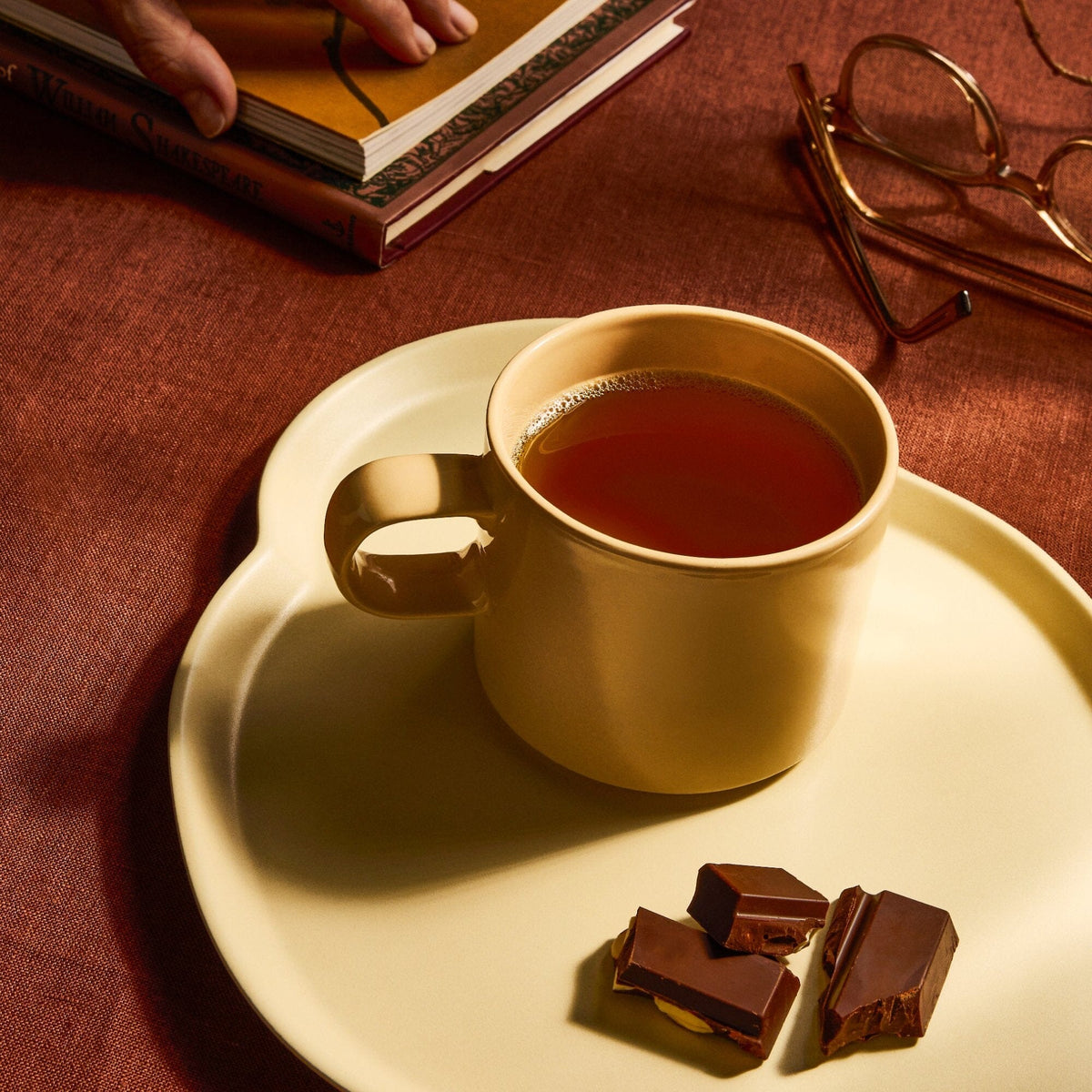 A beige mug of aromatic Rishi Tea & Botanicals peppermint tea sits on a matching plate with chocolate pieces, beside a book, glasses, and a hand resting on a brown tablecloth.
