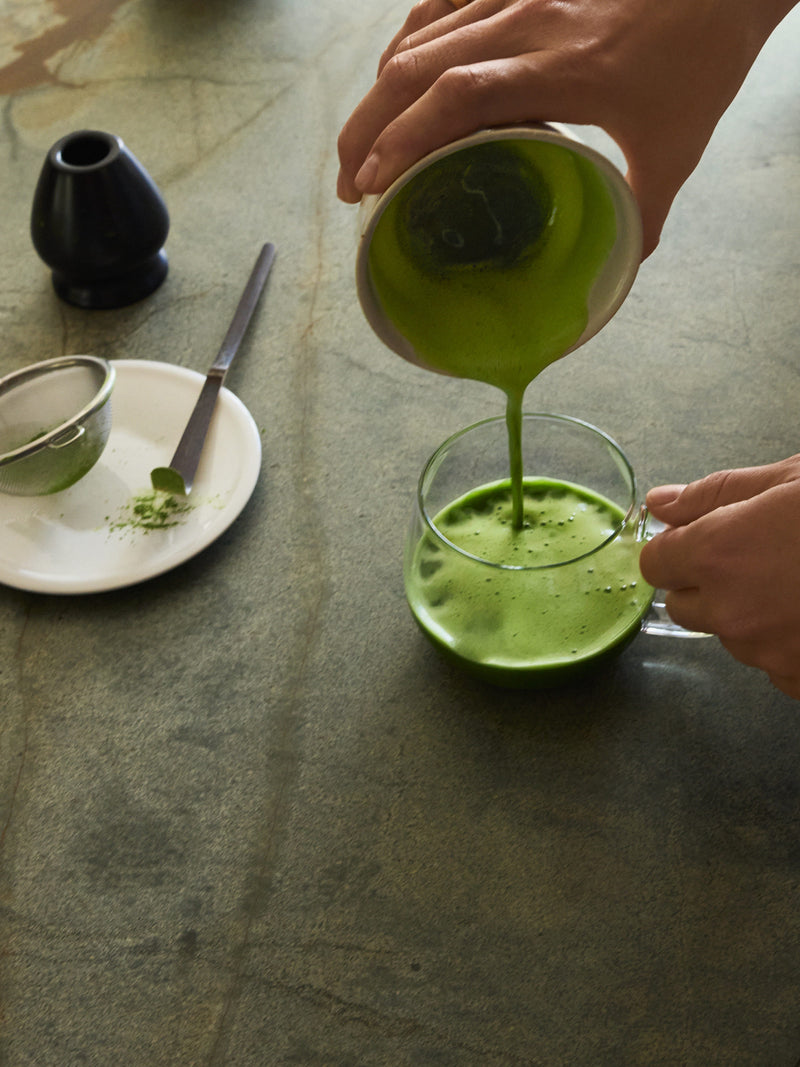 A person's hands pour a vibrant green matcha tea from a small white bowl into a clear glass mug on a textured gray stone surface, with a matcha strainer and small black vessel on a white plate nearby.