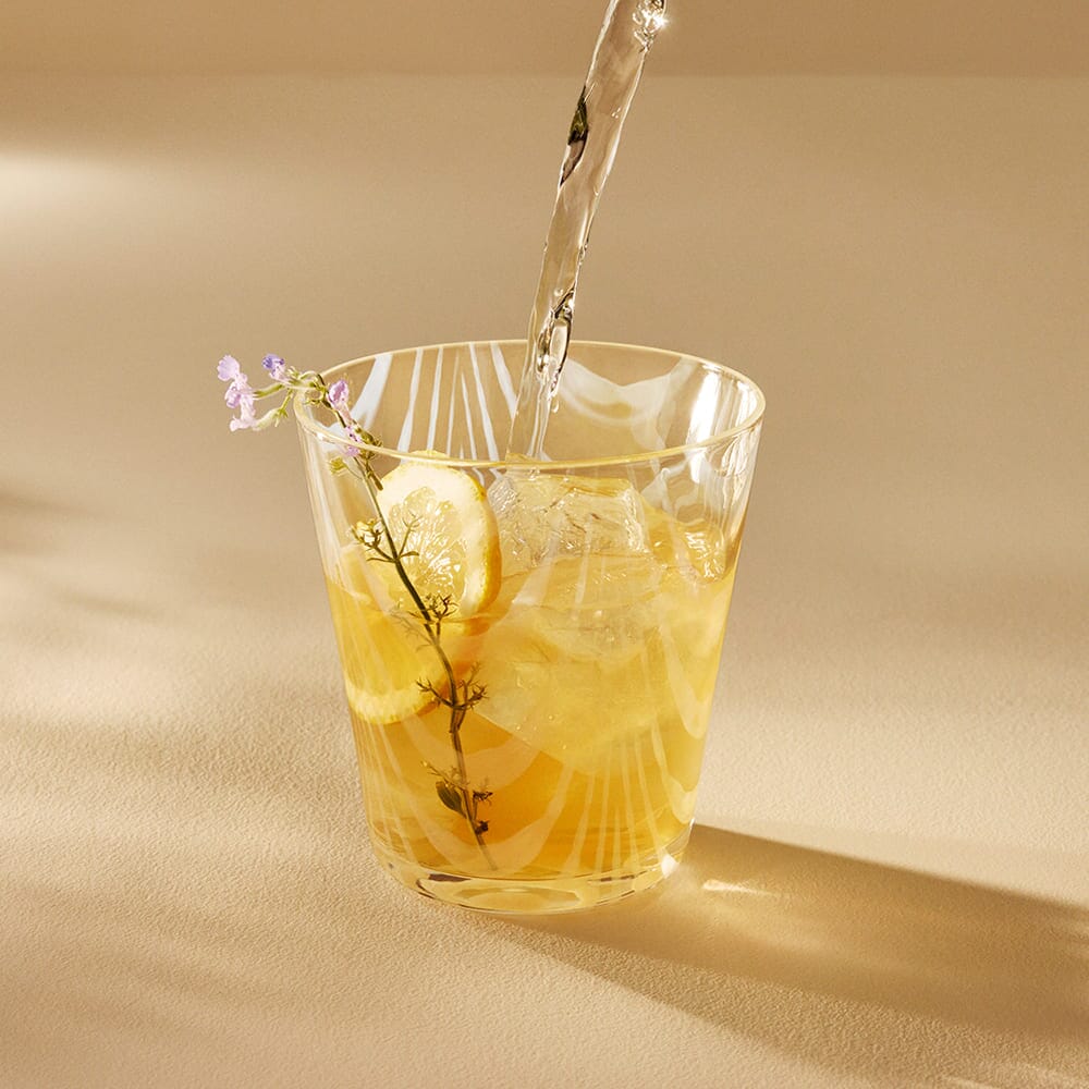 A glass of iced lemon drink from Rishi Tea & Botanicals, garnished with herbs and soothing lavender flowers, is being filled with liquid on a light-colored surface.
