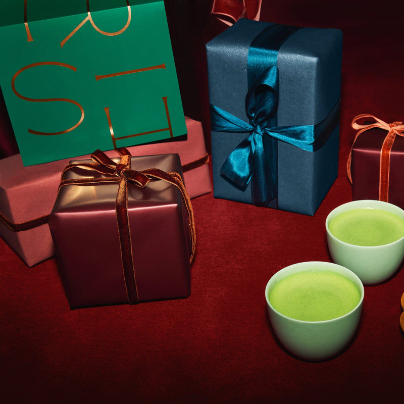 Four wrapped gift boxes and two cups of green matcha tea are arranged on a red surface.