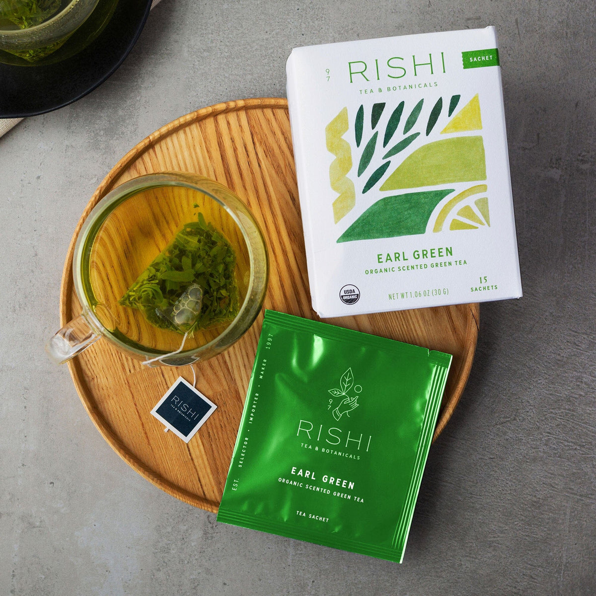 A cup of green tea with a hint of Earl Grey sits alongside a green tea bag, a green sachet, and a box labeled "Earl Green by Rishi Tea & Botanicals" on a round wooden tray.