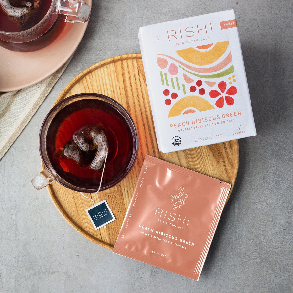 A box of Rishi Tea & Botanicals Peach Hibiscus Green rests elegantly with a tea sachet on a wooden tray, next to a glass cup filled with vibrant red tea.