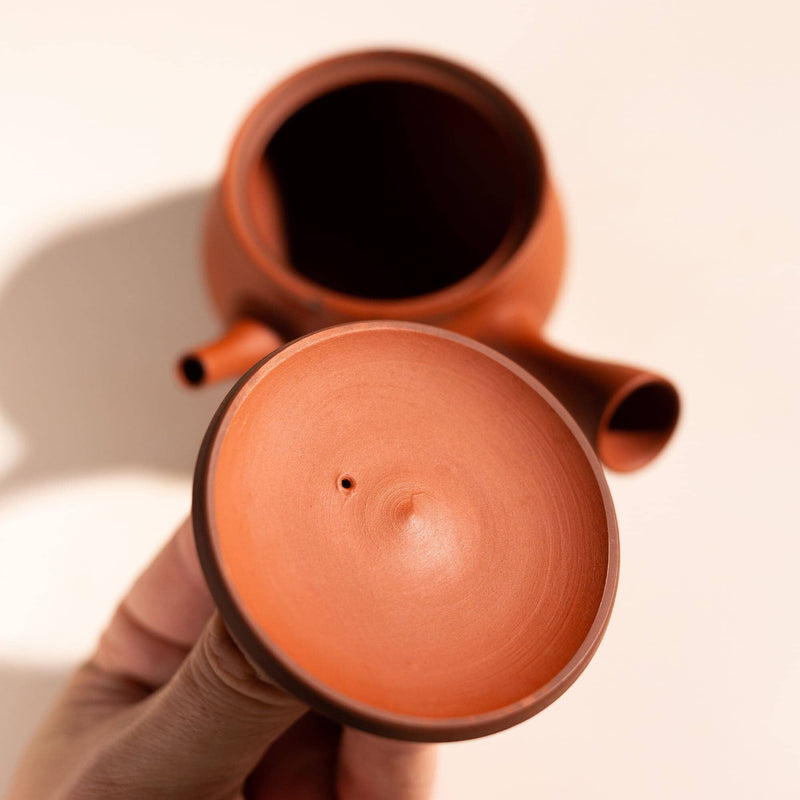 A terracotta-colored clay teapot with its lid removed and held in a hand, photographed against a clean off-white background with a soft shadow.