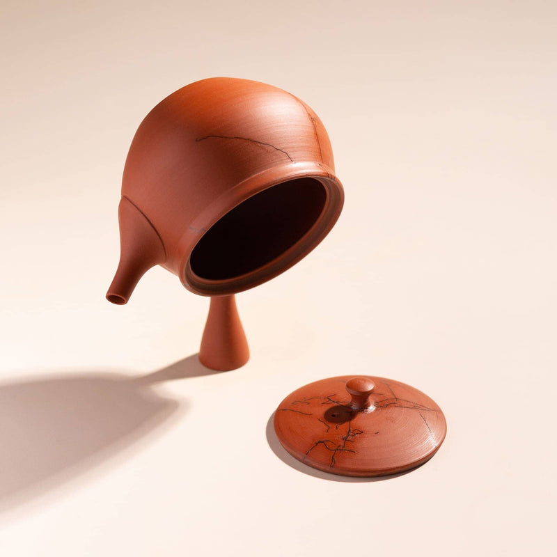A terracotta-colored ceramic teapot with a matching lid, both featuring a natural black crack pattern, displayed on a warm beige background with a soft shadow.